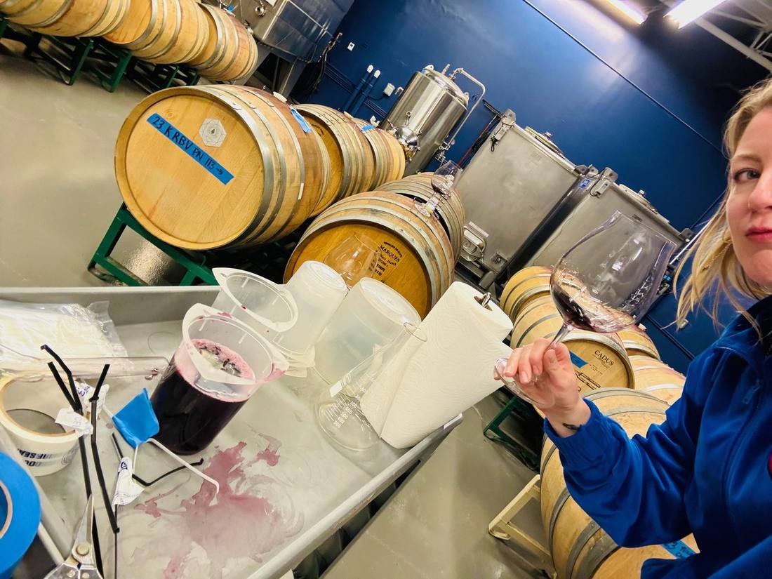 Kendal Montgomery holding up a glass of red wine beside a blending bench of beakers and a flask, barrels behind her